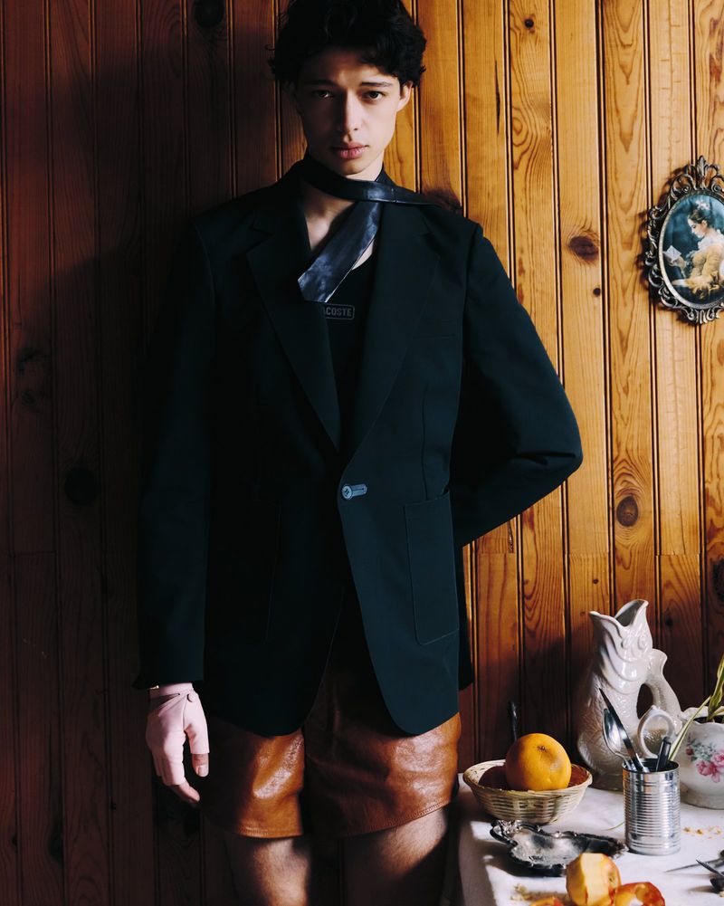 Portrait of a young adult wearing a black jacket, dark tie, and leather shorts, posed against a wood paneled wall with decorative objects.