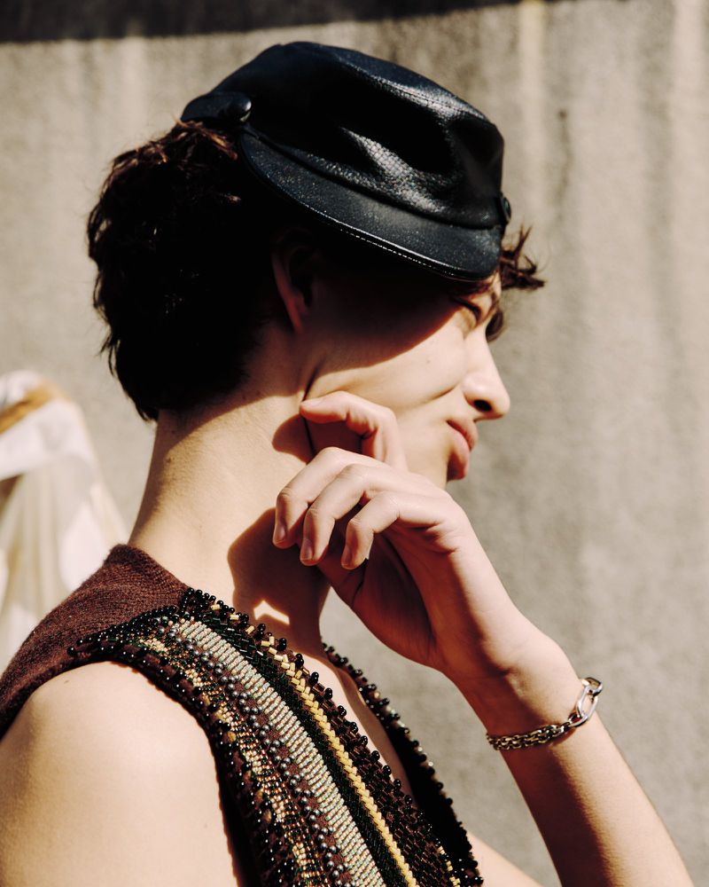 Profile view of a young adult wearing a black cap, a beaded brown vest, and a silver bracelet, set against a neutral backdrop in soft light.
