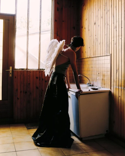 Woman wearing angel wings leans against a sink in a wooden-paneled room with window. Light from the window illuminates the scene.