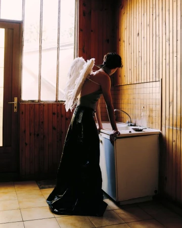 Woman wearing angel wings leans against a sink in a wooden-paneled room with window. Light from the window illuminates the scene.