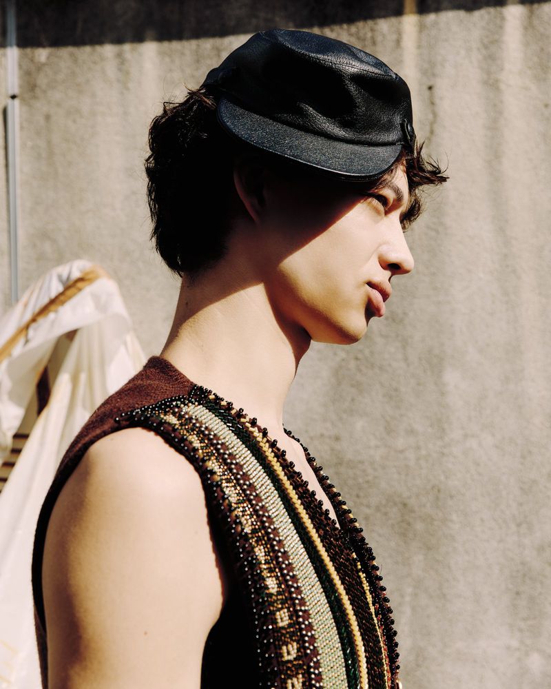 Portrait of a young adult man with curly hair wearing a leather cap and beaded vest against a neutral concrete backdrop.