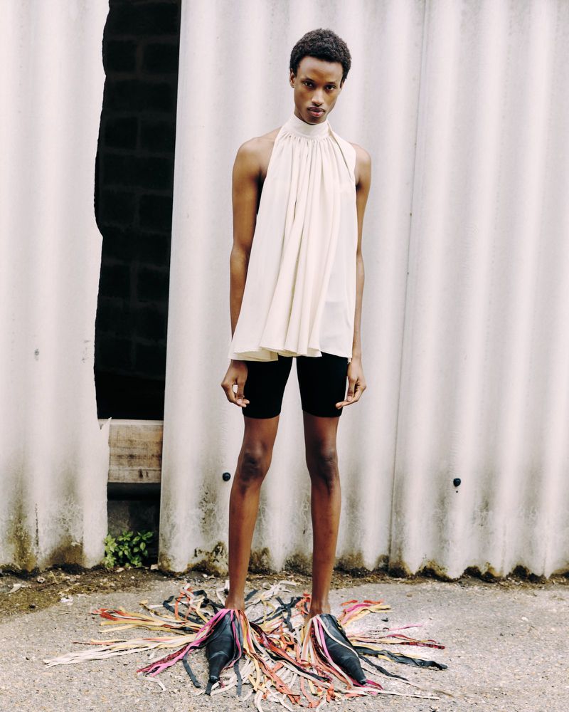 Fashionable person in halter top and black shorts poses outdoors with decorative shoes, showcasing an avant-garde, editorial aesthetic.