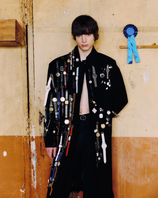 Portrait of a young adult man wearing an open black coat covered in wristwatches, standing in front of a weathered wall with a blue ribbon.