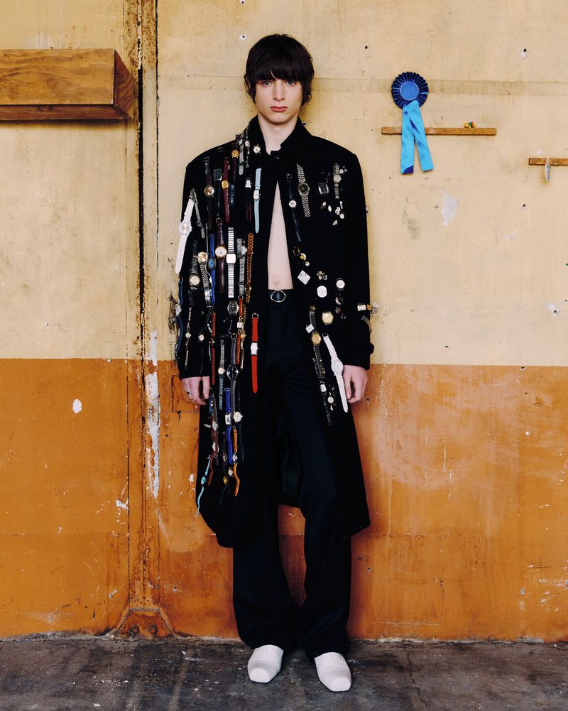 An adult male poses in a decorated black coat, black pants, and white shoes, set against a rustic wall with an attached blue ribbon.