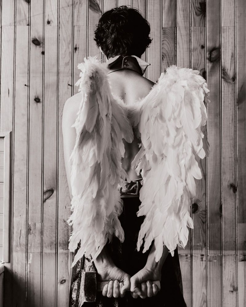 Black and white studio shot from the back of a person wearing large feathered wings and holding hands behind back, standing by wood paneling.