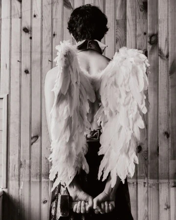 Black and white studio shot from the back of a person wearing large feathered wings and holding hands behind back, standing by wood paneling.