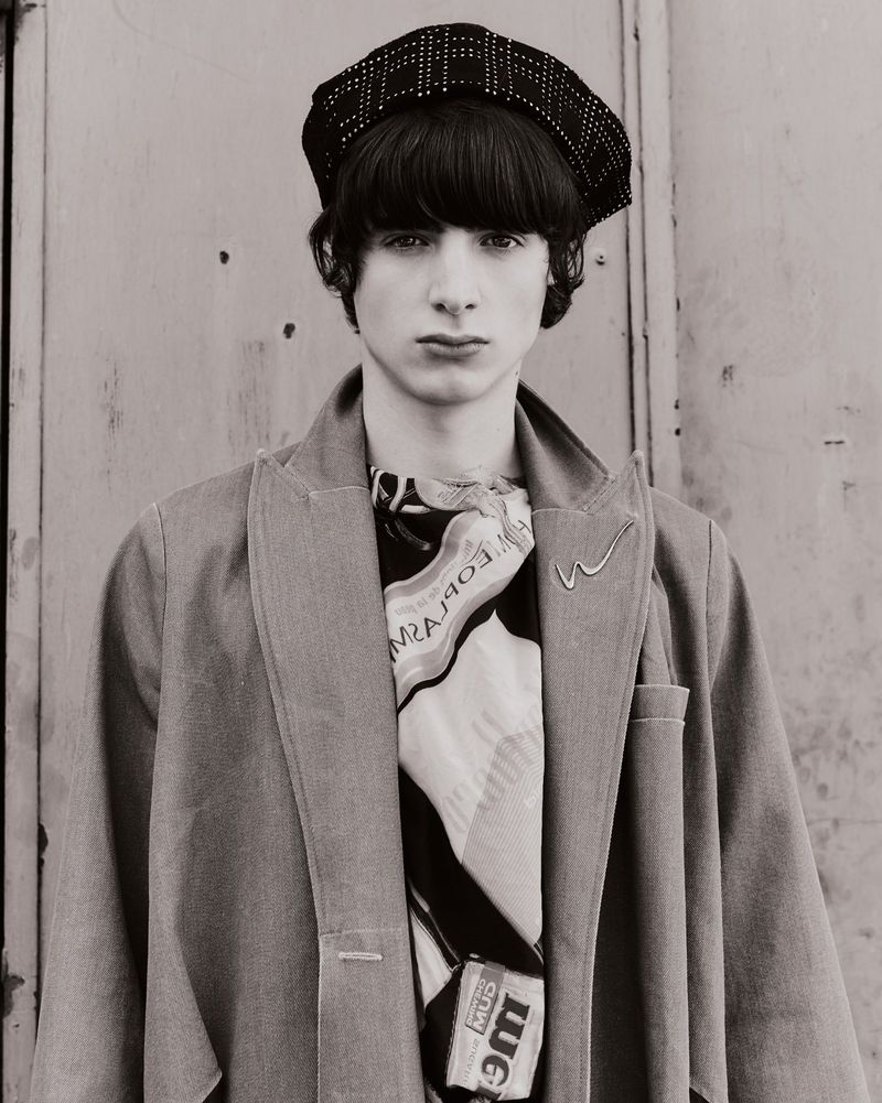 Monochrome portrait of a young man in a patterned hat and coat against a muted backdrop, with a scarf featuring graphic elements.