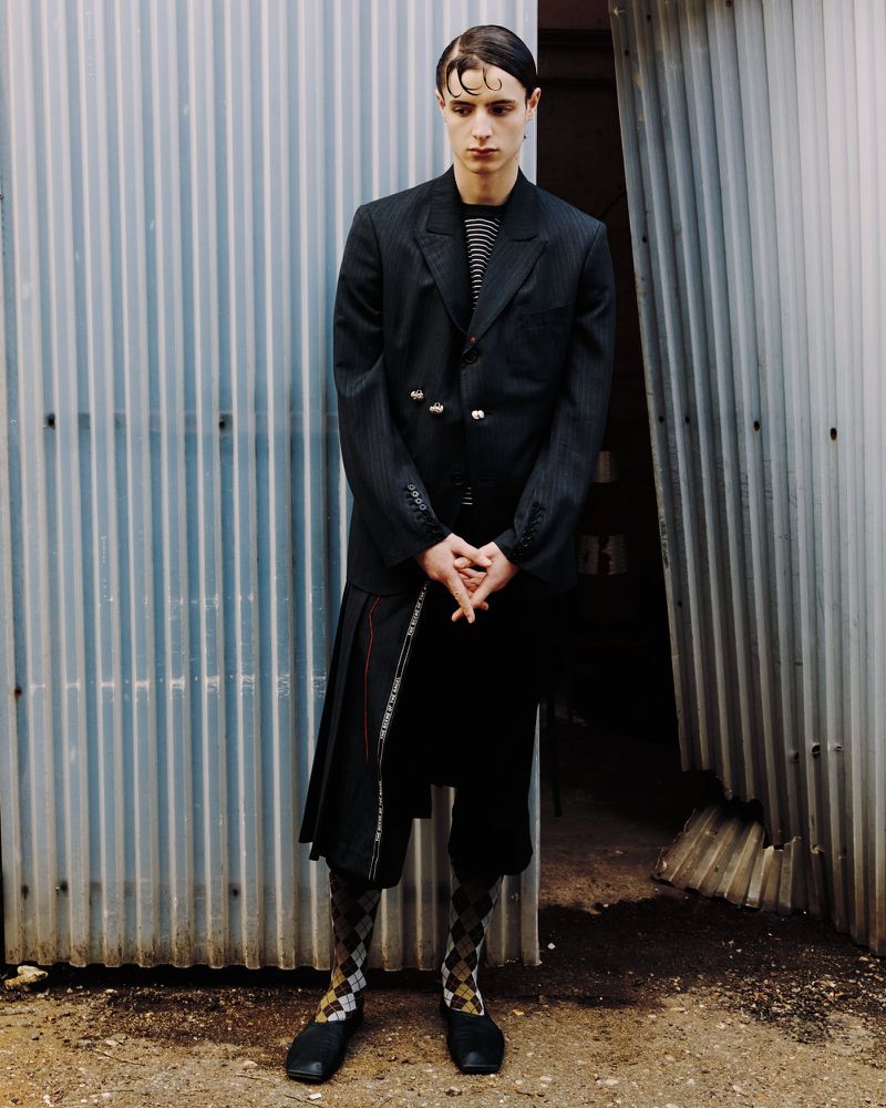 A young man in a black blazer and skirt stands in front of a corrugated metal wall, showcasing a unique fashion ensemble.