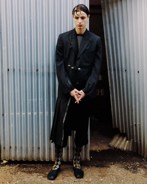 A young man in a black blazer and skirt stands in front of a corrugated metal wall, showcasing a unique fashion ensemble.
