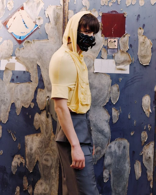 Person in yellow hooded top wearing a black face mask, posing in front of a peeling wall with blue and white layers.