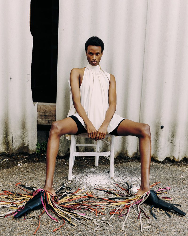Young adult woman sits on a chair outside, wearing a halter dress and black shorts with sculptural heels decorated with colorful tassels.