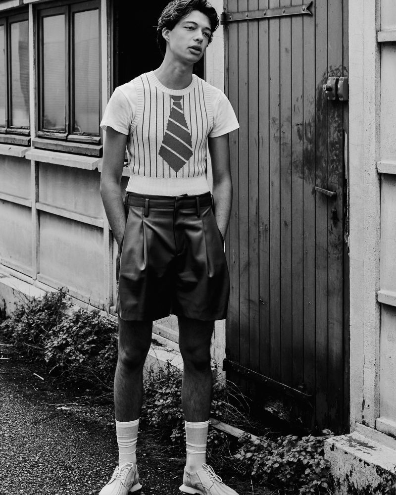 Monochrome portrait of a young man in a striped t-shirt with a tie graphic, paired with leather shorts and white socks, standing outdoors near a door.