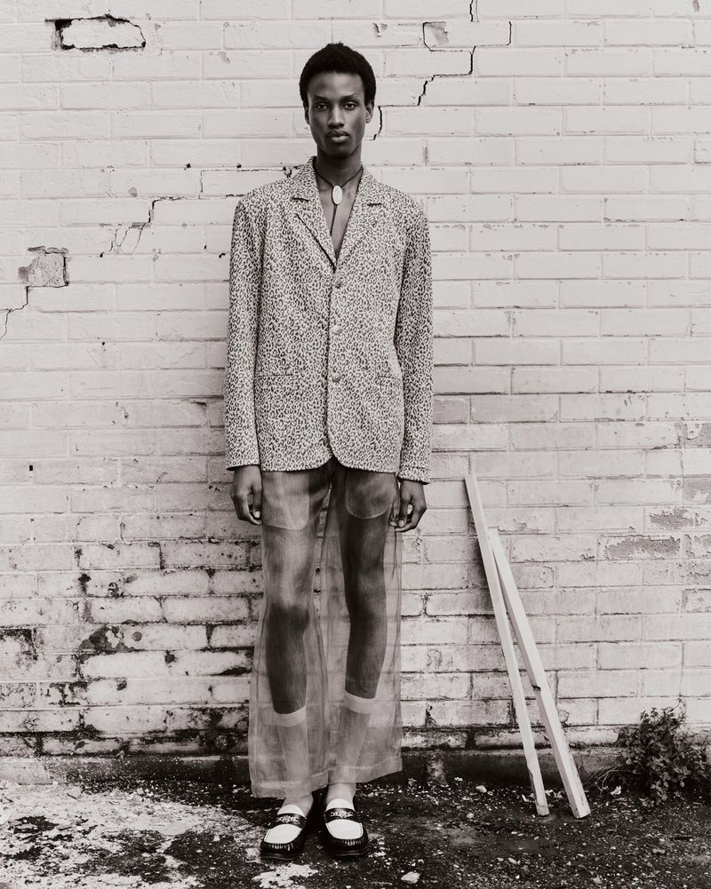 An androgynous fashion model poses in front of a white brick wall wearing a patterned blazer, sheer pants, and white-trimmed black loafers.