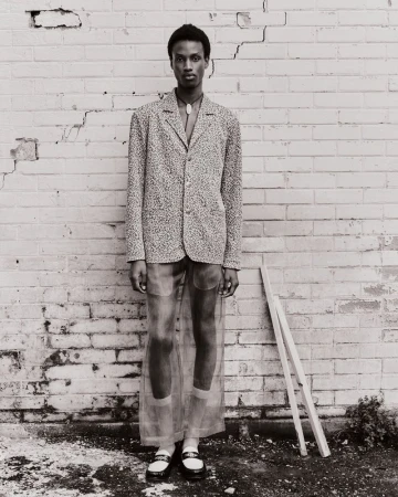 An androgynous fashion model poses in front of a white brick wall wearing a patterned blazer, sheer pants, and white-trimmed black loafers.