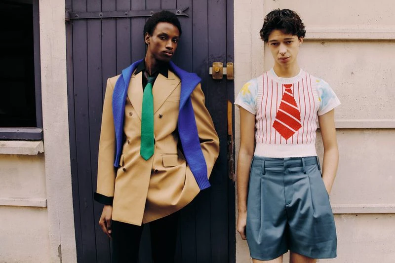 Two people pose in a fashion shot near a blue door. One wears a beige jacket with a green tie, the other a striped shirt and blue shorts.