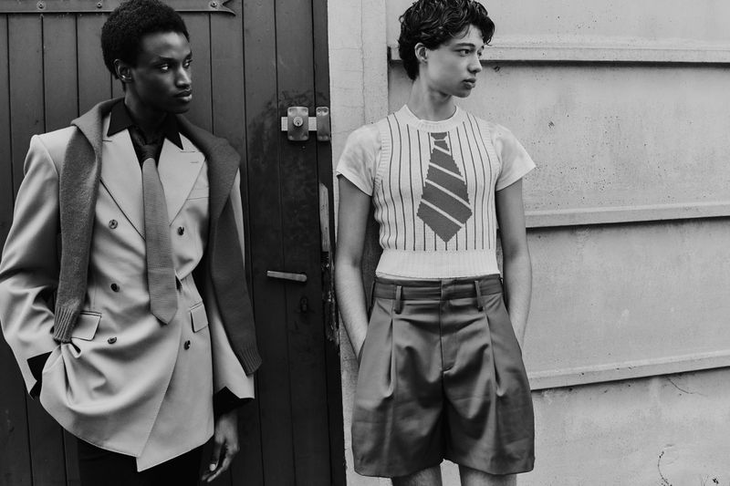 Two stylish young men pose outdoors; one in a double-breasted suit with a scarf, the other in a tie-themed top and shorts.
