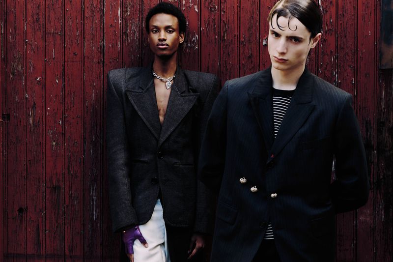 Two dark-haired men pose against a textured red wall in black, structured blazers. One wears a necklace and purple glove. Fashion.