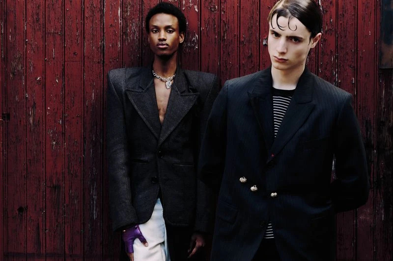Two dark-haired men pose against a textured red wall in black, structured blazers. One wears a necklace and purple glove. Fashion.