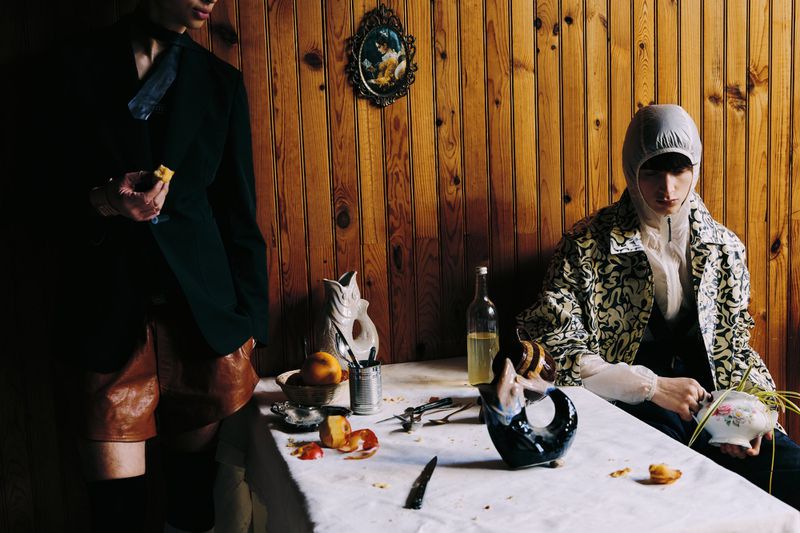 Two people pose indoors near a table covered with fruit and dishes, with a wood-panelled wall in the background.