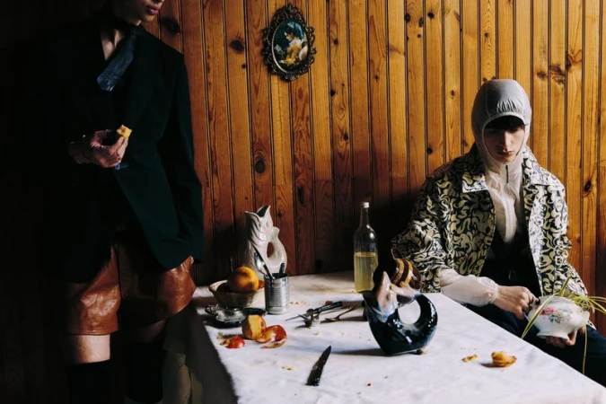 Two people pose indoors near a table covered with fruit and dishes, with a wood-panelled wall in the background.