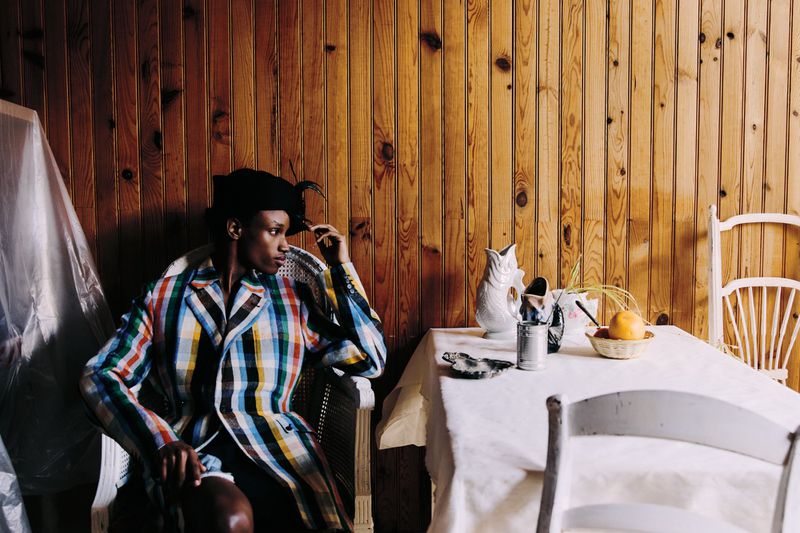Woman in plaid jacket sits at table with fruit, pitcher, and wood paneled wall in background. Indoor fashion lifestyle portrait.