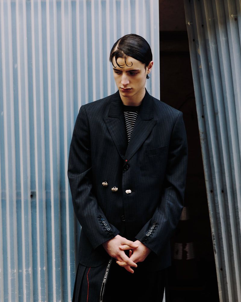 Adult male model in black blazer and striped t-shirt poses against a corrugated metal wall in natural light for a fashion editorial.