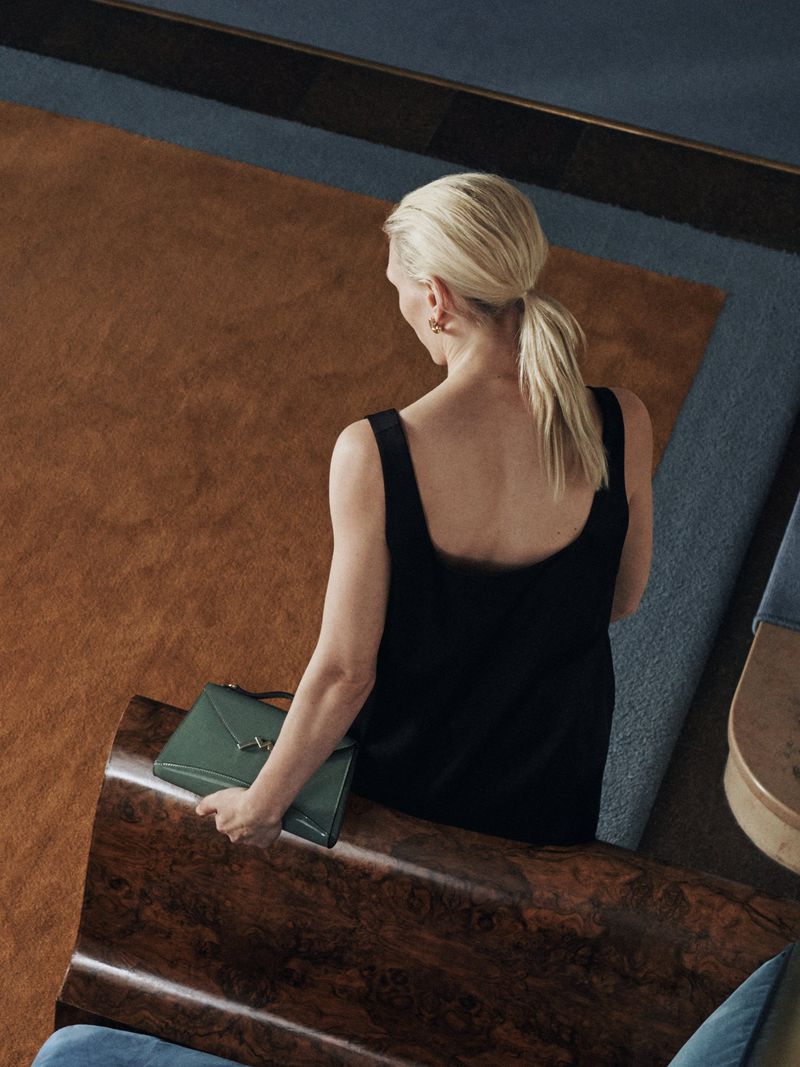 Blonde woman in black dress holds green purse inside a modern room with hardwood floors and a blue accent wall with neutral tones.