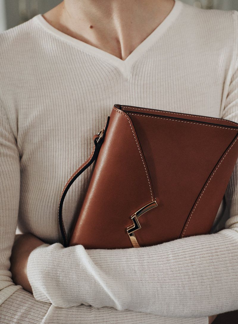 Close-up of a fair-skinned adult female holding a brown leather handbag with a white sweater. Indoor studio lighting.