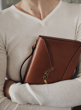 Close-up of a fair-skinned adult female holding a brown leather handbag with a white sweater. Indoor studio lighting.