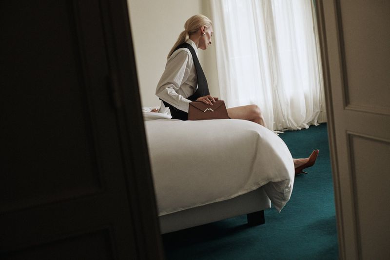 A woman in a white shirt and black vest sits on a bed with a brown purse in a room with a teal carpet and white window curtains.