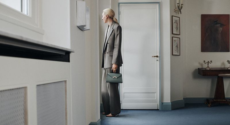 Woman in grey suit holds a green handbag standing indoors near white door and blue carpet, in a fashion or interior setting.