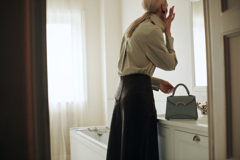 Fashion shot of an adult woman with blonde hair wearing a collared shirt and skirt holding a handbag in a well-lit bathroom setting.