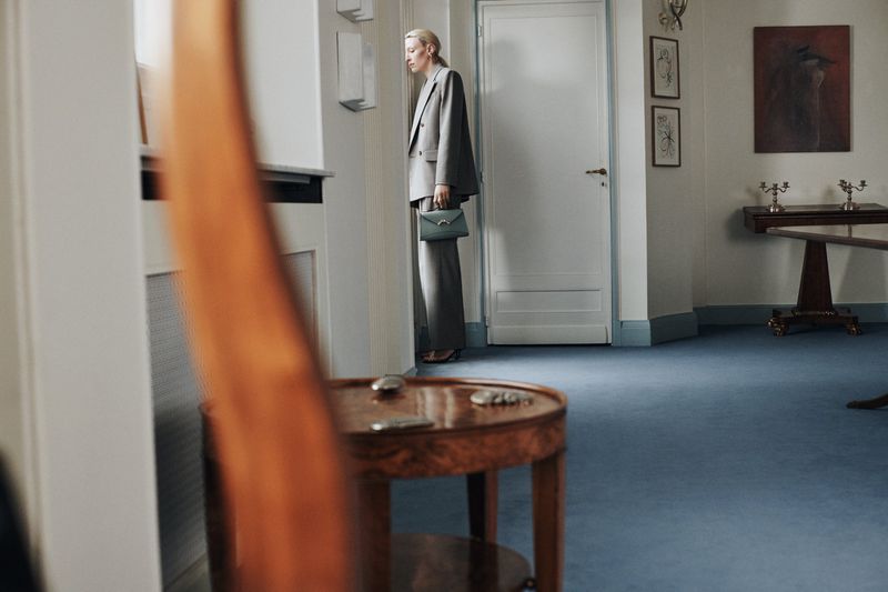 Elegant woman in a grey suit and handbag posing indoors against a white wall, with a blurred table and artwork visible.