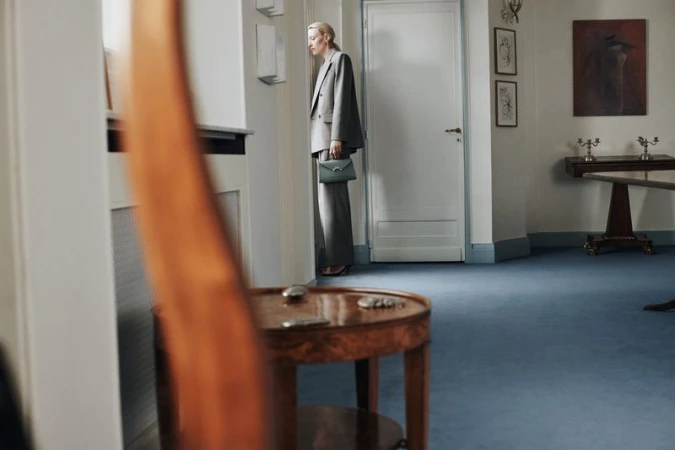 Elegant woman in a grey suit and handbag posing indoors against a white wall, with a blurred table and artwork visible.