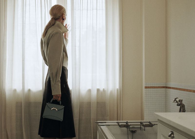 Blonde woman in collared shirt and black skirt holds a blue handbag while looking out sheer window curtains in a bright room.