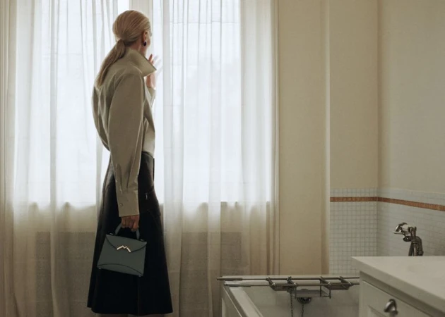 Blonde woman in collared shirt and black skirt holds a blue handbag while looking out sheer window curtains in a bright room.
