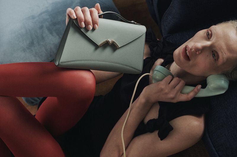 Stylish studio shot featuring a fashionable woman reclining with a modern sage-green handbag, complemented by bright red tights and a vintage rotary phone.