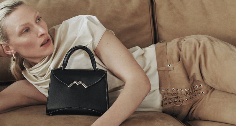 Woman poses with a black leather top-handle handbag on a brown couch, wearing a white shirt and tan pants.