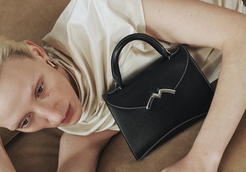 Close-up studio shot of a woman lying down next to a black leather handbag with a silver clasp and white stitching, wearing a tan dress.