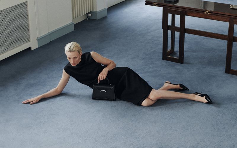 Woman in black dress laying on blue carpet indoors holding a black purse and wearing black high heels. Radiator and table are visible.