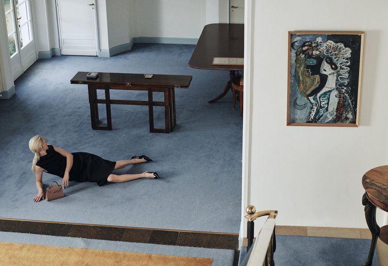 Adult woman in black dress poses on light blue carpet indoors with purse, painting hanging on wall and wooden table in background.