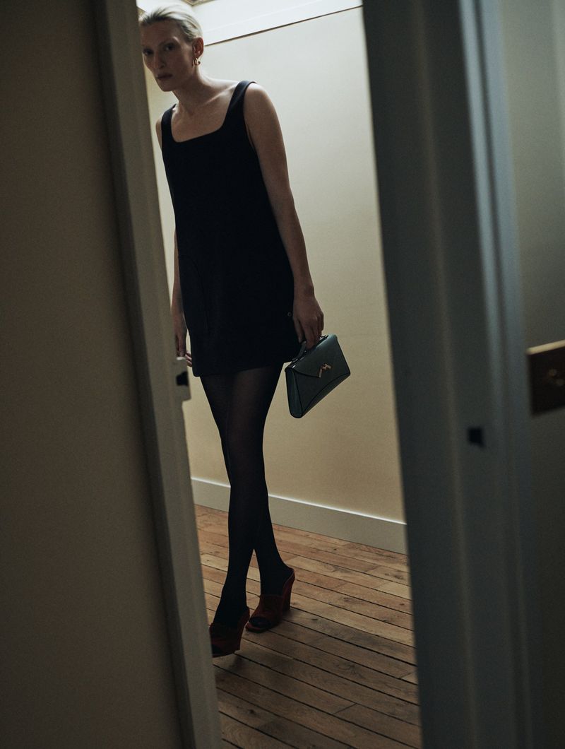 A blonde adult woman stands in a doorway wearing a black dress, tights and heeled shoes, while holding a dark green purse.