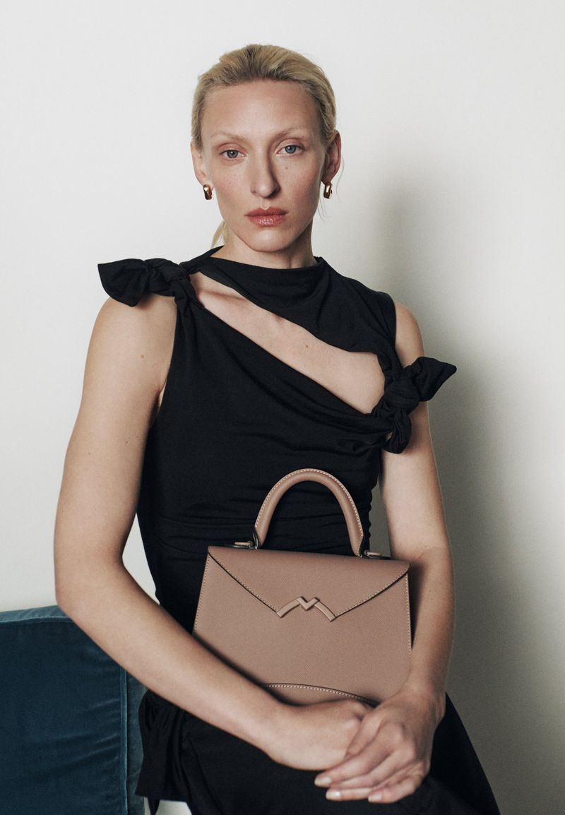 A woman poses in a black dress holding a beige handbag in a studio setting, showcasing fashion, style, and elegance.