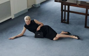 Blonde model in black dress lying on a blue carpet holding a black handbag with dark shoes in an interior