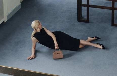 Blonde model in a black dress reclining on a blue carpet holding a leather handbag