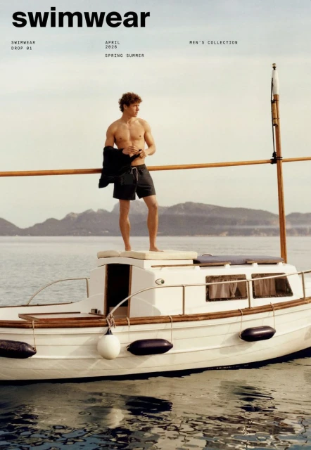 Shirtless man on a white boat with an ocean and island background, advertising swimwear from the Men's Collection for Spring/Summer.