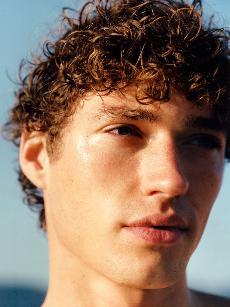 Close-up portrait of a young man with curly brown hair looking off-camera against a clear blue sky background.