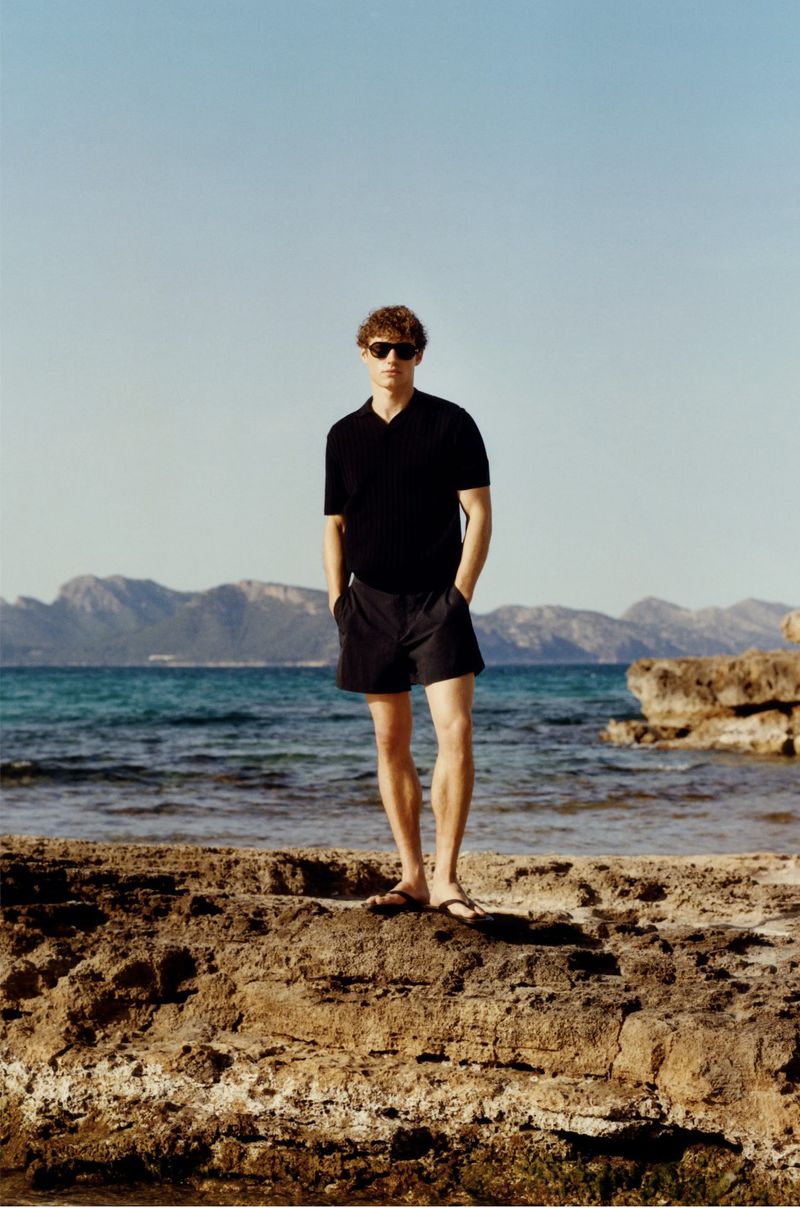 Man in black shirt and shorts stands on rocky beach with mountains and blue sea in background on a sunny day.