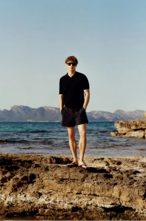 Man in black shirt and shorts stands on rocky beach with mountains and blue sea in background on a sunny day.