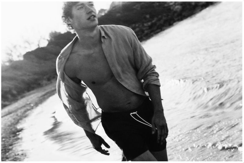 A man with wavy hair wears an unbuttoned shirt and dark swim trunks while walking along a beach shoreline in a black and white photo.